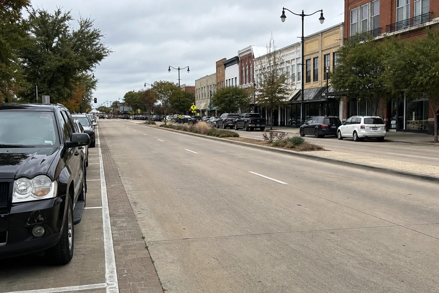 Wide concrete main street lined with brick storefronts, parked cars and young trees under an overcast sky, with a black SUV parked in the foreground