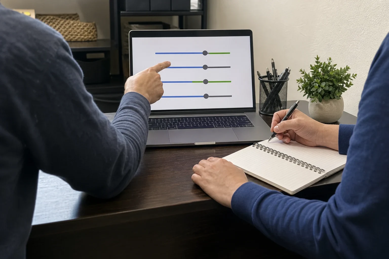 Over-the-shoulder view of one person pointing at adjustable sliders on a laptop while another takes notes in a spiral notebook at a dark wood office desk
