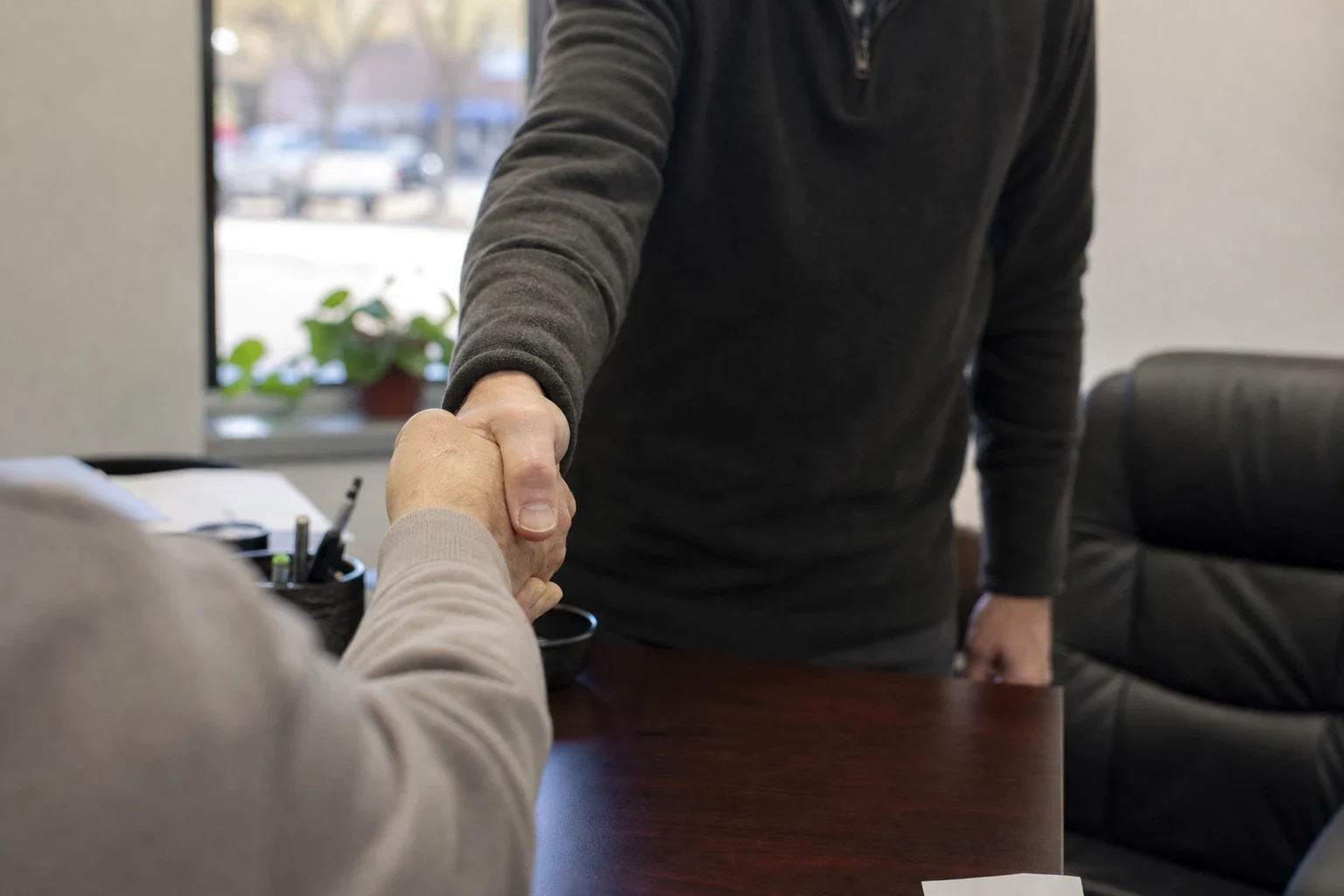 Over-the-shoulder view of two men shaking hands across a wooden office desk, with a leather chair, a potted plant and a window onto a street with parked cars behind.